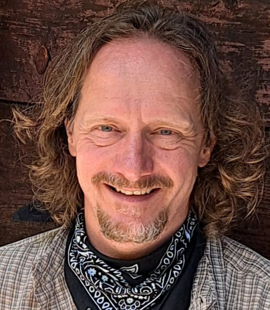 Der (Life-)Coach ist in Portraitaufnahme vor einem  Holzhaus in den Bergen zu sehen.
